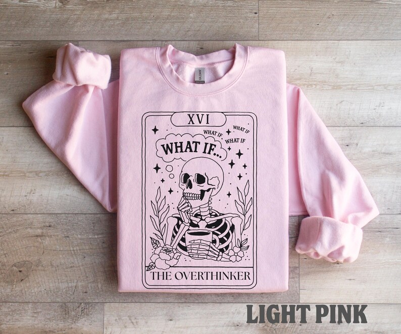 Puede incluir: Sudadera rosa claro con un dise&ntilde;o de carta de tarot en blanco y negro. La carta presenta un esqueleto sosteniendo una taza, con el texto "WHAT IF" y "THE OVERTHINKER". Las mangas est&aacute;n dobladas.