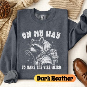 May include: Dark heather grey sweatshirt with a white graphic of a raccoon wearing a hooded jacket and the text "On My Way To Make The Vibe Weird".