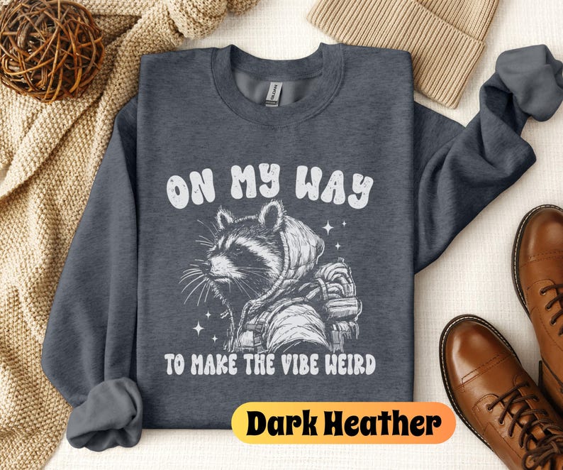May include: Dark heather grey sweatshirt with a white graphic of a raccoon wearing a hooded jacket and the text "On My Way To Make The Vibe Weird".