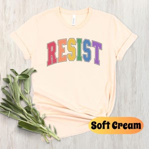 May include: A soft cream colored t-shirt with the word "RESIST" printed in rainbow colors. The word is in a varsity style font.