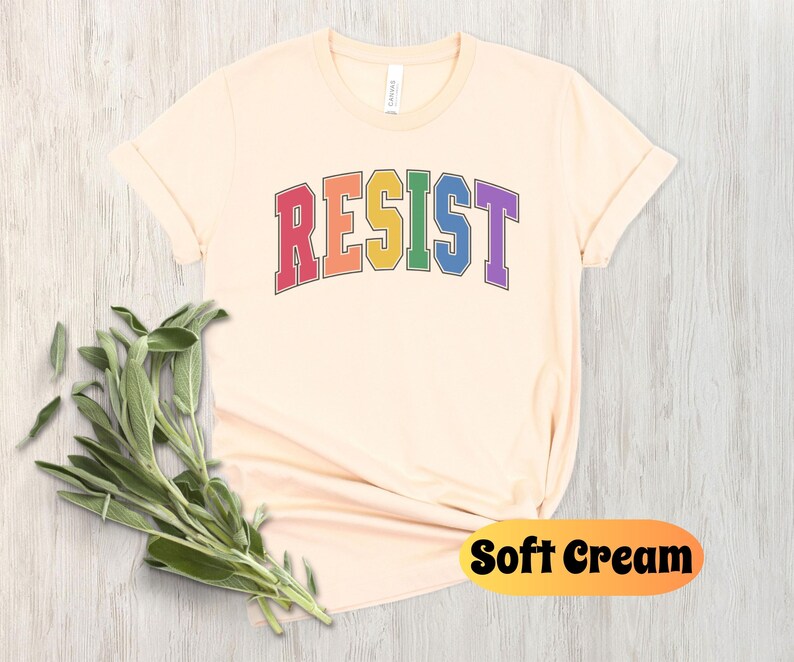 May include: A soft cream colored t-shirt with the word "RESIST" printed in rainbow colors. The word is in a varsity style font.