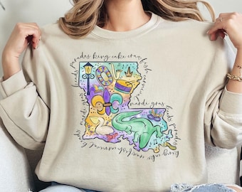 May include: A beige sweatshirt with a graphic print of the state of Louisiana. The graphic includes various symbols and text related to Mardi Gras, including beads, king cake, crawfish, and the fleur-de-lis. The text around the graphic reads "beads parades king cake crawfish carnival mardi gras beads parades".