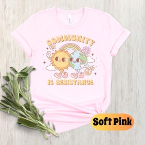 May include: A soft pink t-shirt with a graphic of a sun and earth holding hands. The text "Community is Resistance" is printed below the graphic.