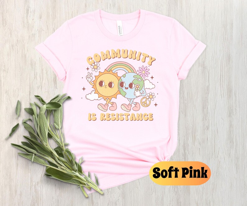 May include: A soft pink t-shirt with a graphic of a sun and earth holding hands. The text "Community is Resistance" is printed below the graphic.