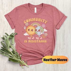 May include: Heather mauve t-shirt with a graphic of a sun and earth holding hands with the text "Community is Resistance".