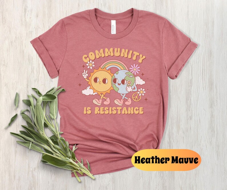 May include: Heather mauve t-shirt with a graphic of a sun and earth holding hands with the text "Community is Resistance".
