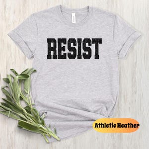 May include: Athletic heather grey t-shirt with the word "RESIST" printed in a distressed black font. The shirt is folded on a light wood surface with a bundle of green leaves. The tag on the shirt reads "Athletic Heather".