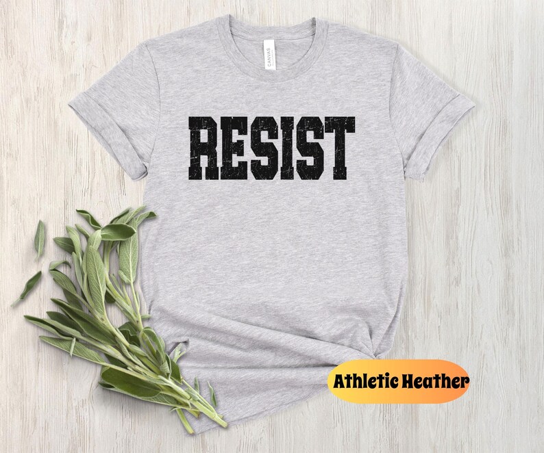 May include: Athletic heather grey t-shirt with the word "RESIST" printed in a distressed black font. The shirt is folded on a light wood surface with a bundle of green leaves. The tag on the shirt reads "Athletic Heather".