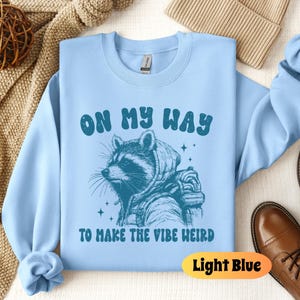 May include: Light blue sweatshirt with a raccoon wearing a hoodie and the text "On My Way To Make The Vibe Weird".