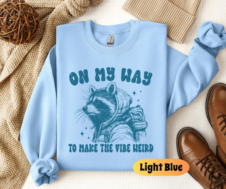 May include: Light blue sweatshirt with a raccoon wearing a hoodie and the text "On My Way To Make The Vibe Weird".