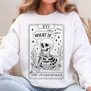 Puede incluir: Sudadera blanca con un dise&ntilde;o de carta de tarot en blanco y negro. La carta representa un esqueleto en posici&oacute;n pensativa, con las palabras "WHAT IF..." y "THE OVERTHINKER". El dise&ntilde;o incluye elementos florales y estrellas.