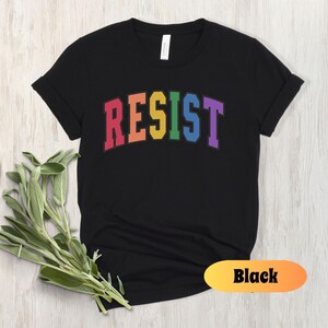 May include: Black t-shirt with the word "RESIST" in rainbow colors. The word is in a collegiate font.