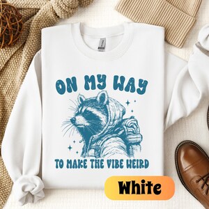 May include: White sweatshirt with a raccoon wearing a backpack and the text "On My Way To Make The Vibe Weird" in blue lettering.