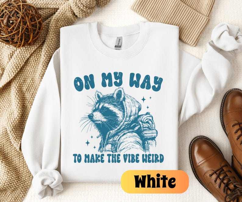 May include: White sweatshirt with a raccoon wearing a backpack and the text "On My Way To Make The Vibe Weird" in blue lettering.