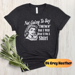 May include: Dark grey heather t-shirt with a retro illustration of a woman holding a cup of coffee. The text "Not Going To Say "I Told You So" But I Will Wear it On A Shirt" is printed on the shirt.
