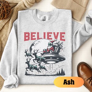 May include: A gray sweatshirt with a graphic of Santa Claus flying in a UFO with reindeer. The text "BELIEVE" is printed above the graphic. The sweatshirt is available in the color Ash.