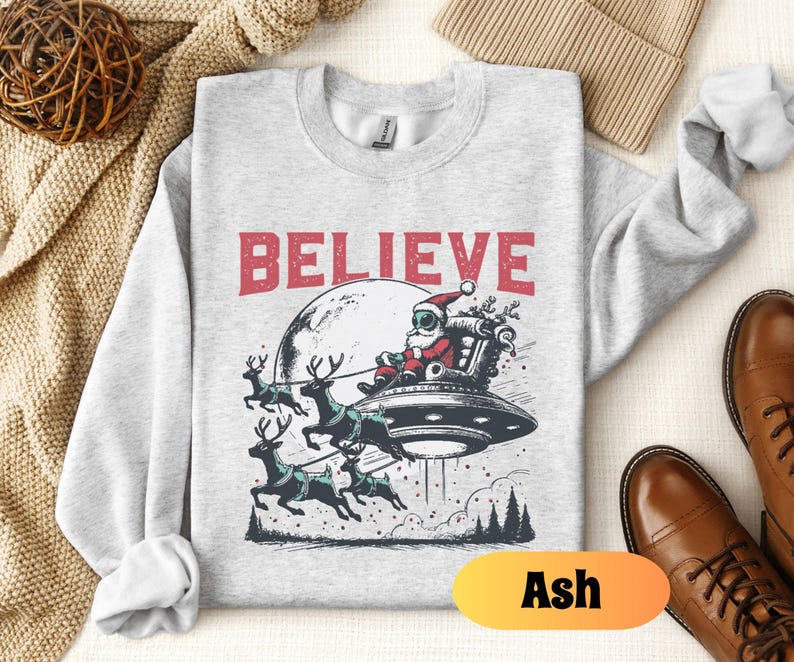 May include: A gray sweatshirt with a graphic of Santa Claus flying in a UFO with reindeer. The text "BELIEVE" is printed above the graphic. The sweatshirt is available in the color Ash.