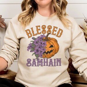 以下が含まれることがあります： クリーム色のスウェットシャツに、オレンジとパープルの文字で「Blessed Samhain」と書かれています。シャツの中央には、パープルの花がついた漫画の南瓜が描かれています。