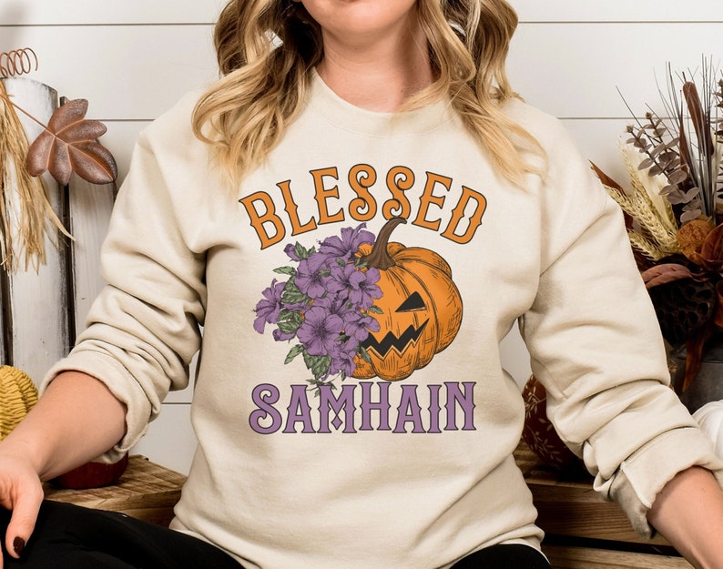 以下が含まれることがあります： クリーム色のスウェットシャツに、オレンジとパープルの文字で「Blessed Samhain」と書かれています。シャツの中央には、パープルの花がついた漫画の南瓜が描かれています。