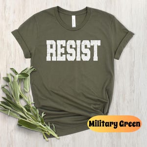 May include: Military green t-shirt with the word "RESIST" printed in white, block letters. The shirt has rolled-up sleeves and a crew neck. A sprig of green leaves is placed near the shirt.