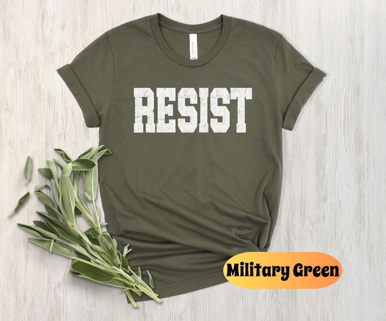 May include: Military green t-shirt with the word "RESIST" printed in white, block letters. The shirt has rolled-up sleeves and a crew neck. A sprig of green leaves is placed near the shirt.