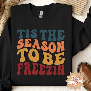 May include: Black sweatshirt with a retro-style graphic that reads "Tis the season to be freezin". The text is in a rainbow of colors, including red, orange, yellow, green, and blue.