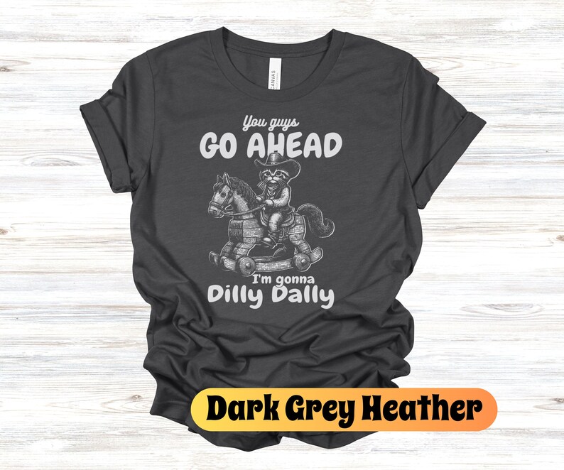 May include: Dark grey heather t-shirt with a white graphic of a raccoon riding a rocking horse. The text "You guys go ahead I'm gonna dilly dally" is printed on the shirt.