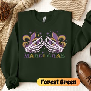 May include: Forest green sweatshirt with a Mardi Gras design featuring two skeletal hands holding fleur-de-lis. The text "Mardi Gras" is printed in yellow and green.