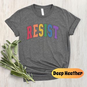 May include: A dark heather grey t-shirt with the word "RESIST" printed in rainbow colors. The word is in a bold, blocky font.