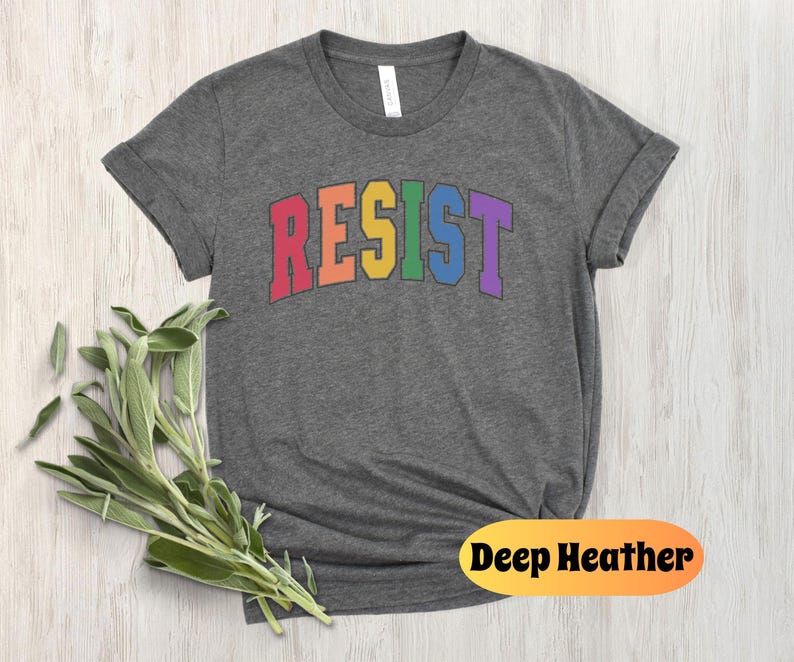 May include: A dark heather grey t-shirt with the word "RESIST" printed in rainbow colors. The word is in a bold, blocky font.