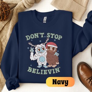 May include: Navy blue sweatshirt with a graphic of two cartoon characters, a yeti and a sasquatch, with the text "Don't Stop Believin'" and the word "Navy" in a yellow box.