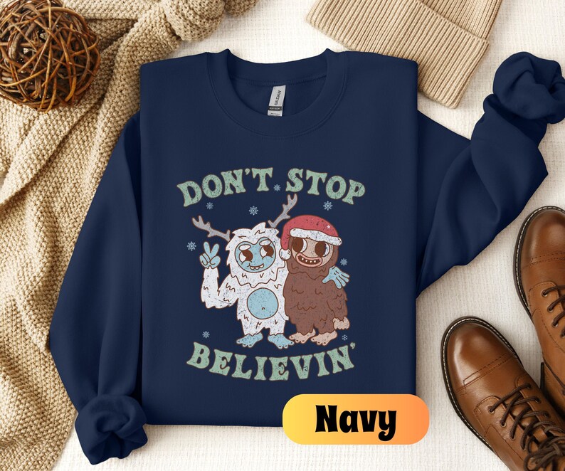 May include: Navy blue sweatshirt with a graphic of two cartoon characters, a yeti and a sasquatch, with the text "Don't Stop Believin'" and the word "Navy" in a yellow box.