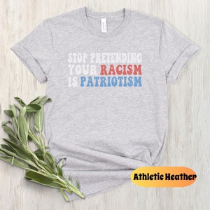 May include: Gray t-shirt with the text "STOP PRETENDING YOUR RACISM IS PATRIOTISM" in red, white, and blue letters. The shirt is on a wood background.