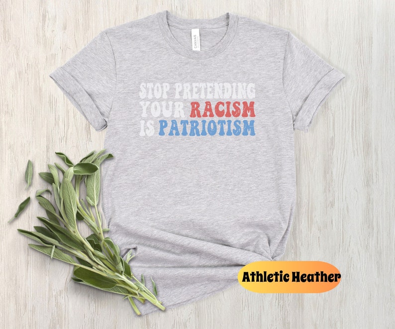 May include: Gray t-shirt with the text "STOP PRETENDING YOUR RACISM IS PATRIOTISM" in red, white, and blue letters. The shirt is on a wood background.