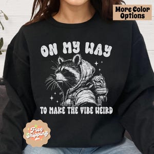 May include: Black sweatshirt with a white graphic of a raccoon wearing a hoodie and backpack. The text "On My Way To Make The Vibe Weird" is printed below the raccoon.