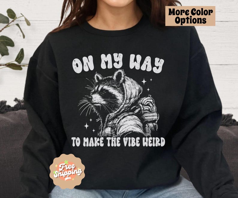 May include: Black sweatshirt with a white graphic of a raccoon wearing a hoodie and backpack. The text "On My Way To Make The Vibe Weird" is printed below the raccoon.