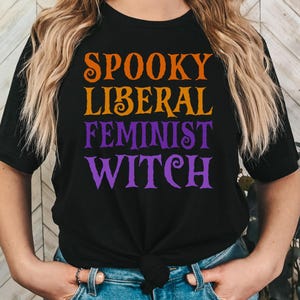 May include: A black t-shirt with the text "Spooky Liberal Feminist Witch" in orange and purple lettering.
