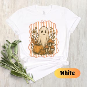 May include: White t-shirt featuring a smiling ghost illustration with a pumpkin, mushrooms, and fall foliage. The design is framed by a peach-colored striped border. The word "White" is displayed in a yellow and orange oval at the bottom.