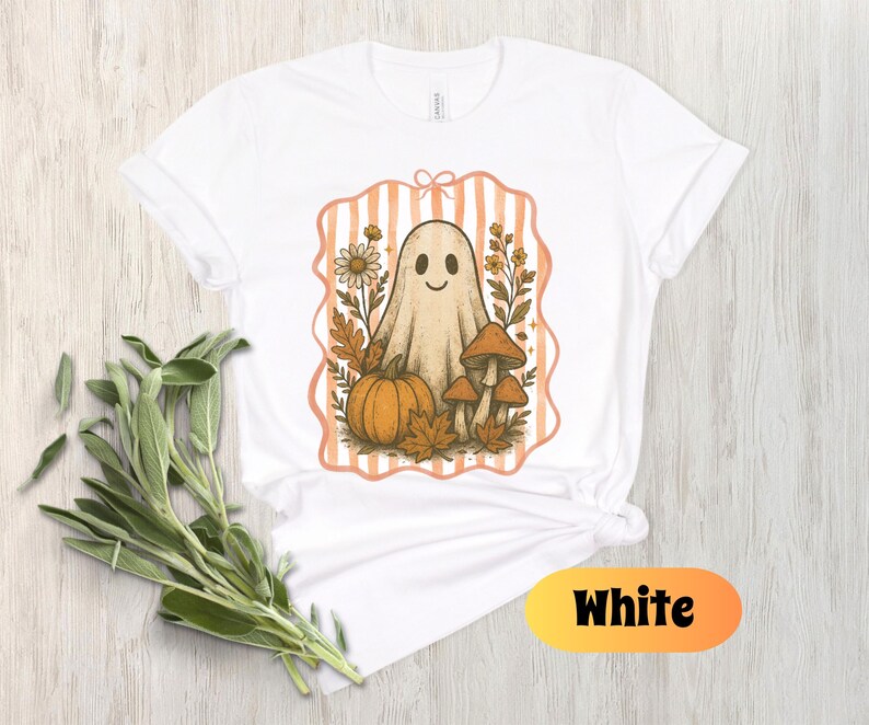 May include: White t-shirt featuring a smiling ghost illustration with a pumpkin, mushrooms, and fall foliage. The design is framed by a peach-colored striped border. The word "White" is displayed in a yellow and orange oval at the bottom.