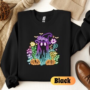 K&ouml;nnte beinhalten: Schwarzes Sweatshirt mit einem neon-gr&uuml;nen, violetten und rosafarbenen Grafikdruck eines Geistes, der einen Hexenhut tr&auml;gt und von Blumen und K&uuml;rbissen umgeben ist.