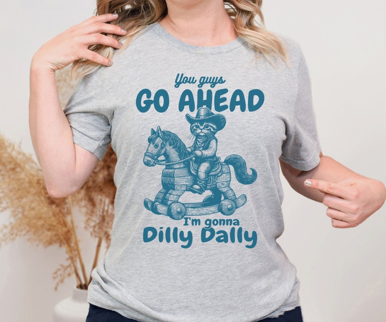 May include: Gray t-shirt with a blue graphic of a cat wearing a cowboy hat and riding a rocking horse. The text "You guys go ahead I'm gonna dilly dally" is printed on the shirt.