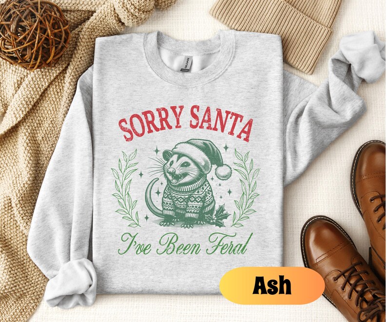 May include: A gray sweatshirt with the text "Sorry Santa I've Been Feral" in red and green. A cartoon possum wearing a Santa hat and a sweater is in the center of the design. The word "Ash" is written in orange on a white background.