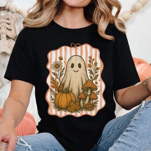 May include: Black t-shirt featuring a whimsical Halloween design. The graphic includes a friendly ghost, pumpkin, mushrooms, and fall foliage within a striped frame. The design is in shades of orange, beige, and brown.