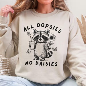 May include: Beige sweatshirt with a black and white graphic of a raccoon holding a sunflower and a drink. The text "ALL OOPSIES NO DAISIES" is printed above and below the raccoon illustration. Butterflies are also included in the design.