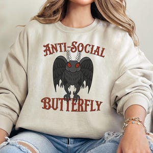 May include: A beige sweatshirt with the text "Anti-Social Butterfly" in red lettering. A black and red moth with red eyes is centered on the sweatshirt.