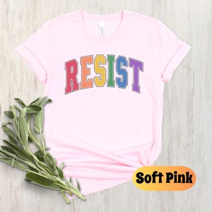 May include: A soft pink t-shirt with the word "RESIST" printed in rainbow colors. The word is in a collegiate font style.