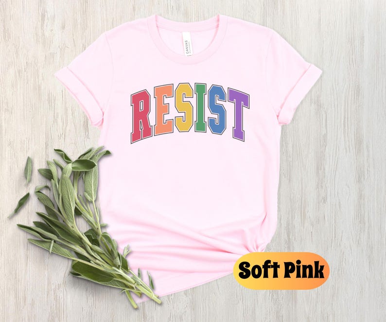 May include: A soft pink t-shirt with the word "RESIST" printed in rainbow colors. The word is in a collegiate font style.