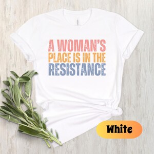 May include: White t-shirt with the text "A Woman's Place Is In The Resistance" in pink, orange, and blue. The shirt is styled with a knot at the waist and is accompanied by a sprig of green leaves. The word "White" is displayed in a yellow and orange oval.