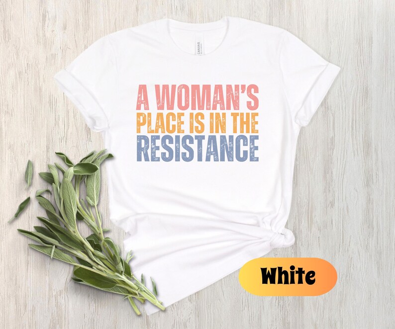 May include: White t-shirt with the text "A Woman's Place Is In The Resistance" in pink, orange, and blue. The shirt is styled with a knot at the waist and is accompanied by a sprig of green leaves. The word "White" is displayed in a yellow and orange oval.