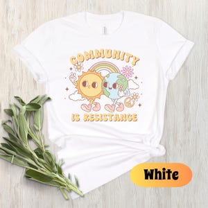 May include: White t-shirt with a graphic of a sun and earth holding hands with the text "Community is Resistance".